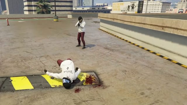 Player in a white hoodie and helmet lying face down on a yellow and black speed bump beside a pool of blood, while a second player aims a pistol from across a rooftop car park.