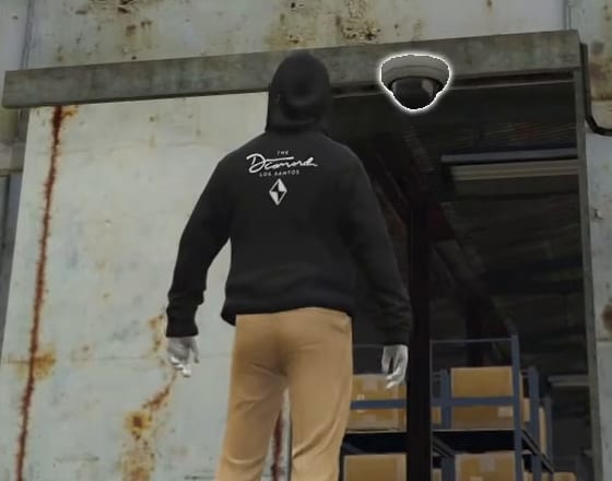 Hooded player seen from behind, looking up at a dome camera outlined in white above a warehouse doorway.