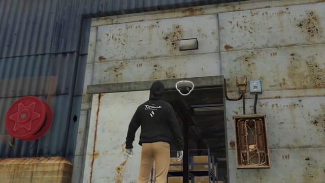 Hooded player looking up at a dome camera, outlined in white, on the lintel above a warehouse doorway, between a red hose reel and a fuse box.