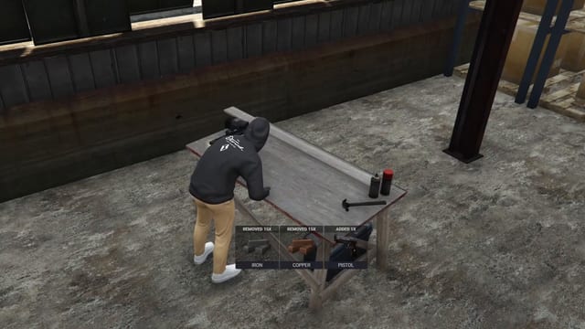 Player leaning over a wooden table with a hammer and two spray cans in a warehouse, above three item notifications: Removed 15x Iron, Removed 15x Copper and Added 1x Pistol.