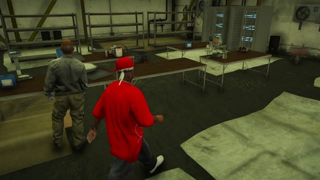 Player in a red shirt and cap walking past a worker in a drug lab with metal shelves, two tall cabinets lit blue, an AC unit and tables of equipment and glassware.