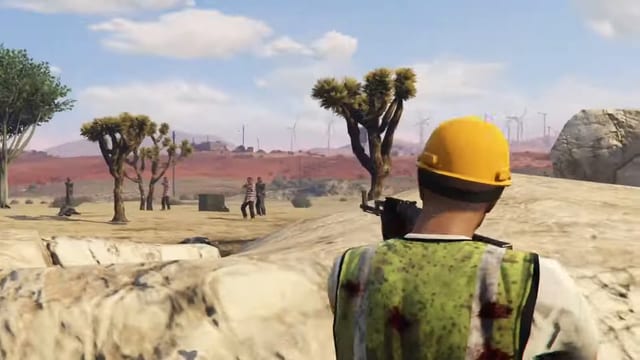 Player in a hard hat and hi-vis vest aiming a rifle over a rock at four men standing around a crate between desert trees, with a fifth man on the ground.