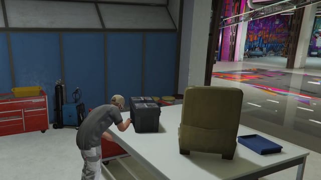 Mechanic bent over a white workbench with a black toolbox on it, beside a red tool chest and a welder, with the painted Bennys garage floor behind.