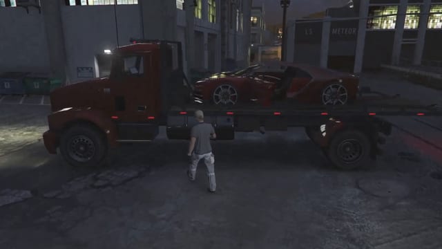 Mechanic walking beside a red flatbed truck at night with a red sports car loaded on its bed, in a street between warehouses.